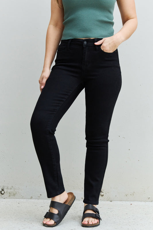A woman embracing modern comfort with Judy Blue Kenya Full Size Mid Rise Slim Fit Jeans. The jeans feature a mid-rise design and a slim fit, creating a sleek and stylish look. Made with quality denim, they guarantee both comfort and durability. Their classic design adds a touch of timeless charm, suitable for any occasion. The woman pairs them with a casual tee for a relaxed vibe or a button-up shirt for a more polished style